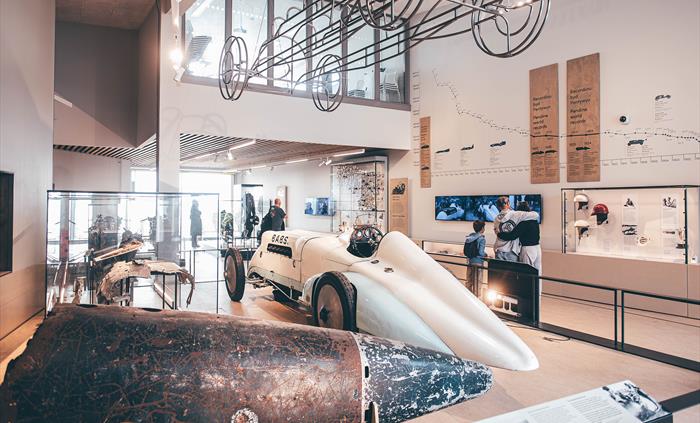 Wide angle view of the main museum gallery. A metal tail from the Bluebird car is in the foreground, with the Babs car in the middle ground. A steel f